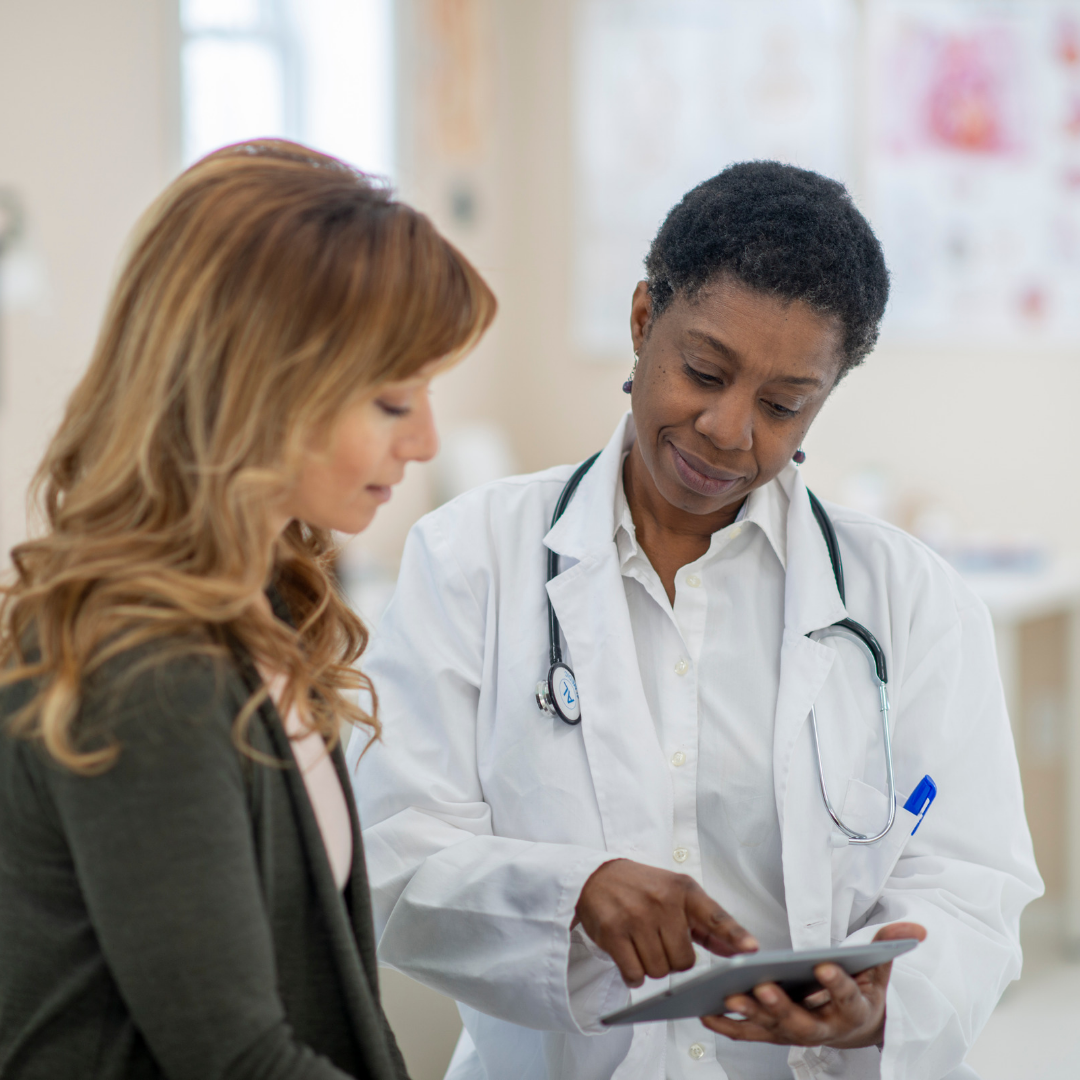A woman doctor in a white coat with a stethoscope around her neck pointing at an tablet and a patient is next to her looking at the tablet
