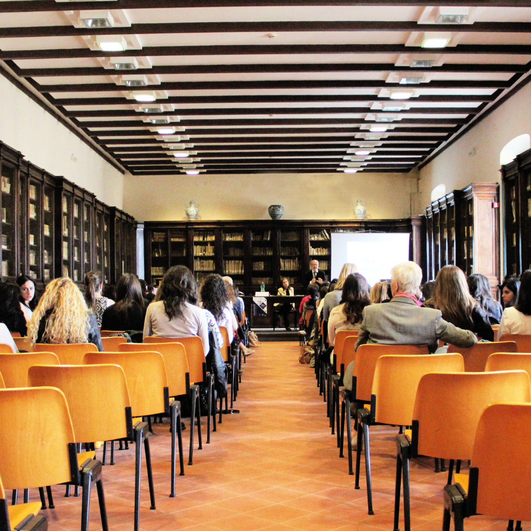Rows of chairs in a conference room with people seated and a projection screen at the front where a man is presenting